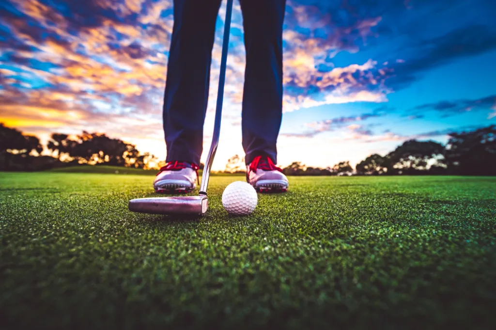 Golf ball on the green with stunning sunset sky and anonymous golfer wearing golf shoes and holding club