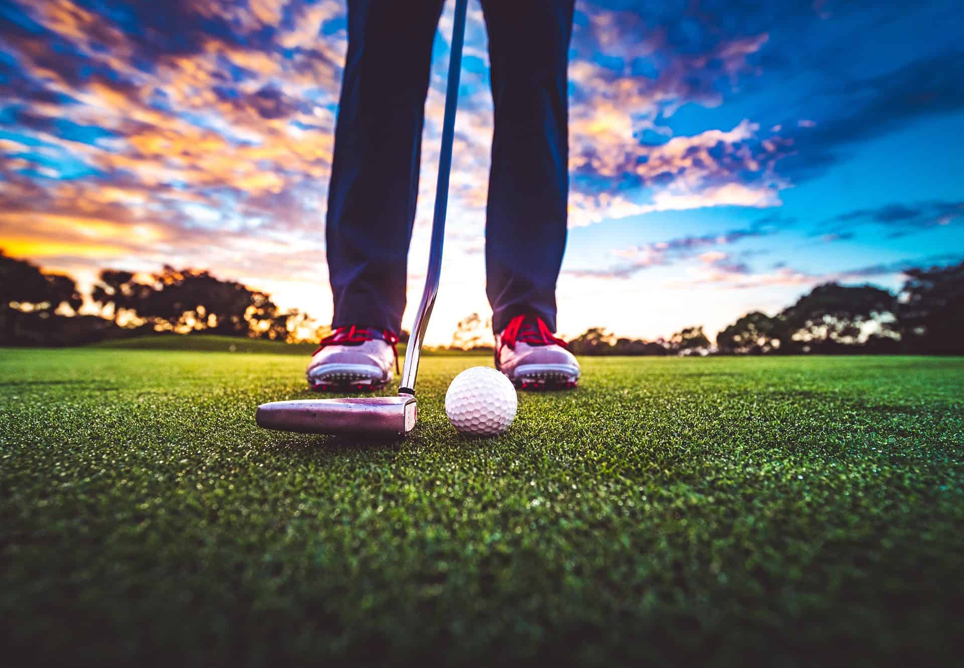 Golf ball on the green with stunning sunset sky and anonymous golfer wearing golf shoes and holding club